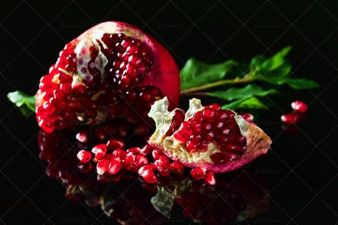 عکس با کیفیت برگ های سبز در کنار انار نصف شده با دانه های یاقوتی انار در پس زمینه مشکی
