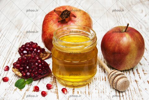 عکس با کیفیت ظرف عسل در کنار انار و اسلایس انار و سیب بر روی میز سفید