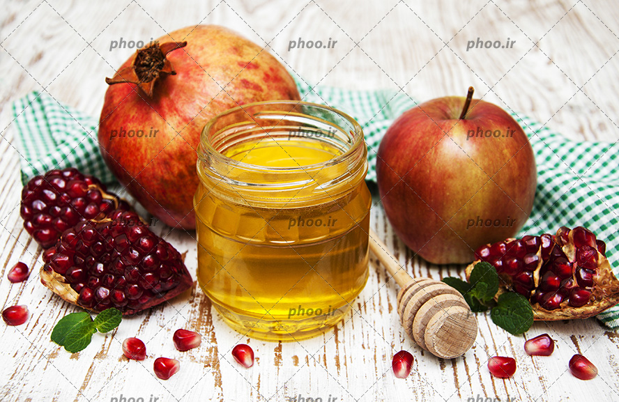 عکس با کیفیت پارچه سبز چهارخونه در پشت سیب و انار و اسلایس های انار در کنار ظرف عسل و قاشق عسل خوری در کنار ظرف