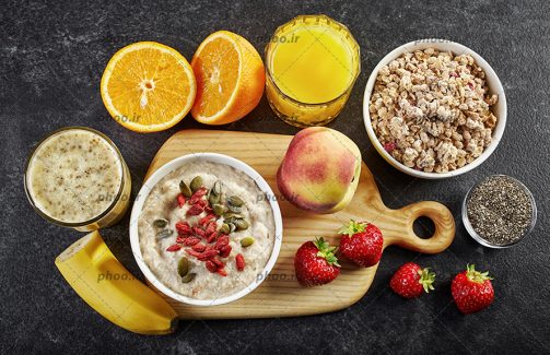 عکس با کیفیت غلات صبحانه در داخل ظرف در کنار آبمیوه پرتقال و موز و توت فرنگی و هلو قرار گرفته بر روی تخته چوبی