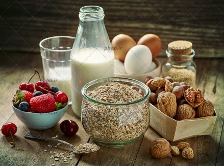 عکس با کیفیت بطری و لیوان شیر در کنار غلات صبحانه و ظرف تخم مرغ و ظرف توت فرنگی ها و گردو ها در کنار یکدیگر
