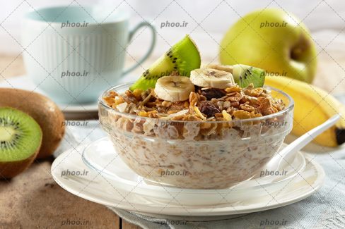 عکس با کیفیت اوتمیل رژیمی میوه ای و مغزیجات و میوه بر روی آن در ظرف شیشه ای در کنار موز و کیوی