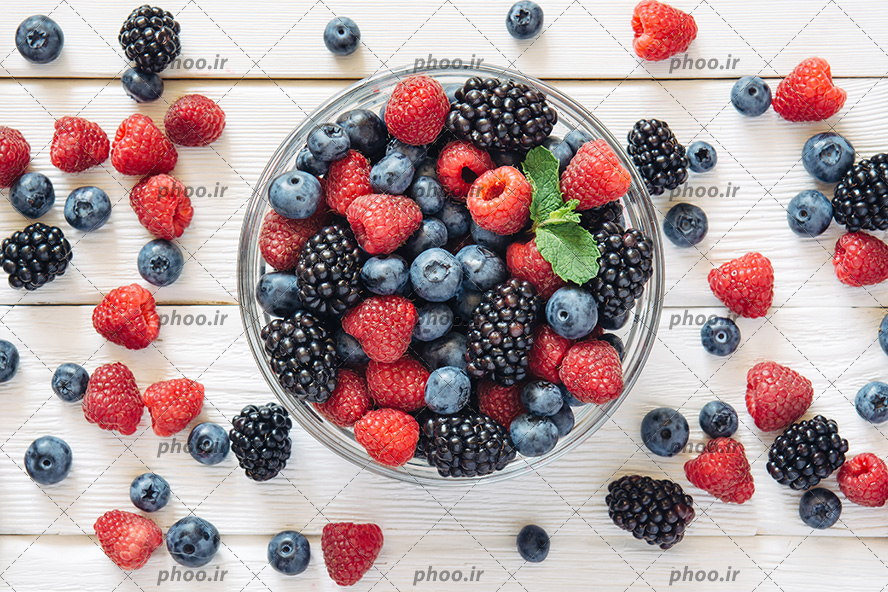 عکس با کیفیت شاتوت و بلوبری و تمشک در ظرف شیشه ای و در کنار ظرف بر روی میز چوبی سفید