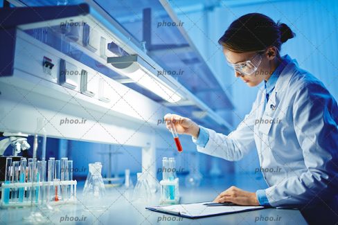 عکس با کیفیت زن با موهای بسته و لباس سفید و عینک در حال نگاه کردن به شیشه های آزمایش و نوشتن گزارش