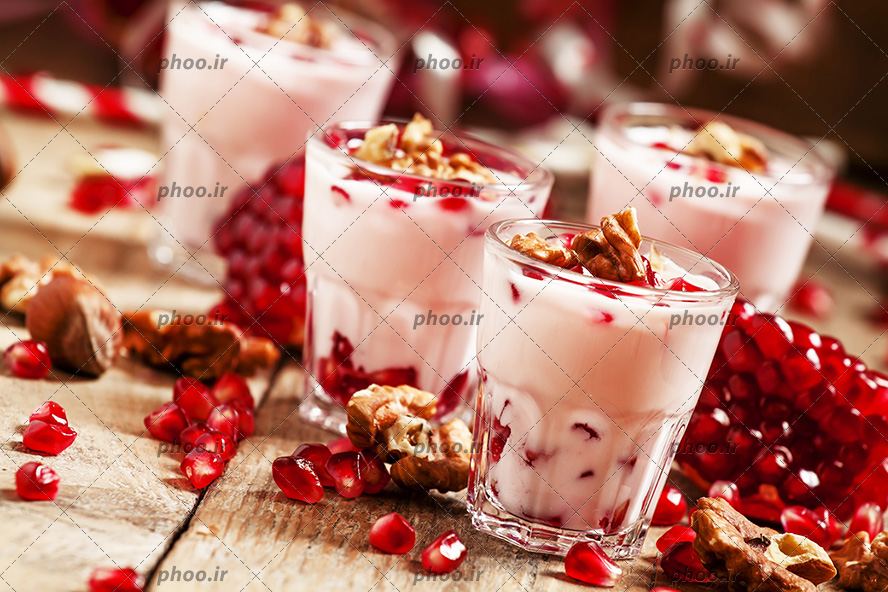 عکس با کیفیت ظروف شیشه ای حاوی دسر های اناری بر روی میز چوبی و اسلایس های انار و گردو در کنار ظروف