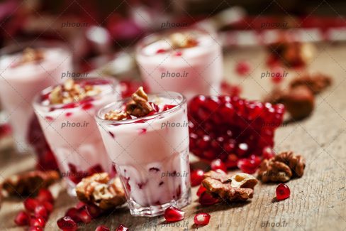عکس با کیفیت گردو ها در کنار دانه ها و اسلایس های انار و ظروف دسر و آنها بر روی میز چوبی