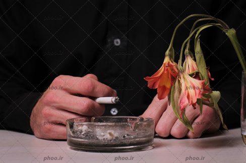 عکس با کیفیت مرد با لباس مشکی در حال سیگار کشیدن و در حال تکاندن خاکستر سیگار در زیر سیگاری