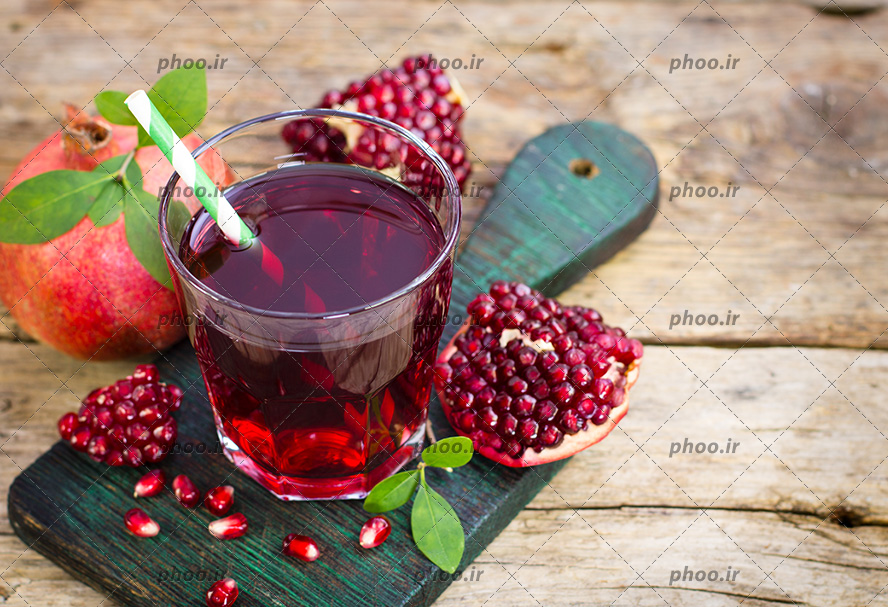 عکس با کیفیت تکه ای انار در کنار لیوان آبمیوه انار و قرار گرفته بر روی تخته چوب فیروزه ای
