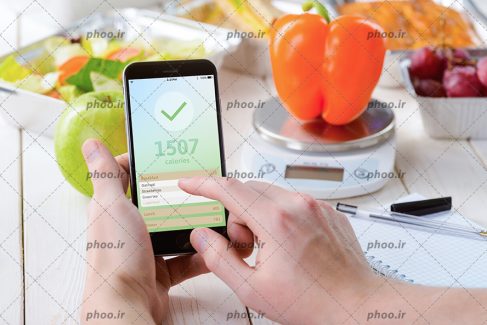عکس تعدادی میوه و سبزیجات روی ترازوی آشپزخانه و موبایلی که میزان کالری آنها را نشان می دهد