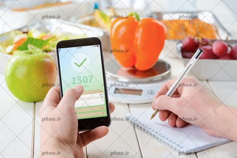 عکس یک ترازو آشپزخانه به همراه تعدادی میوه و سبزیجات و یک قلم و کاغذ برای نوشتن میزان کالری هر کدام از آنها از طریق نرم افزار محاسبه کالری