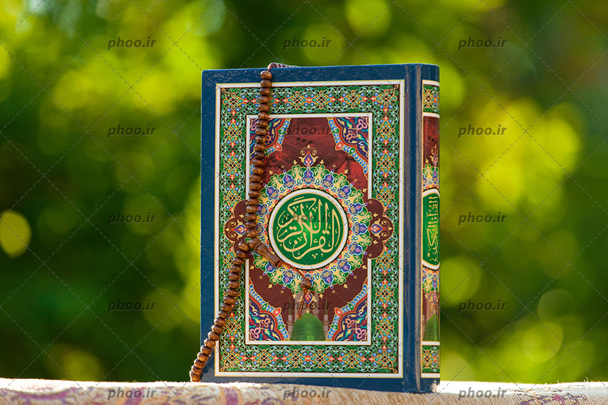 عکس یک کتاب قرآن کریم با جلد رنگی رنگی که یک تسبیح روی آن است و پشت آن درختان زیادی قرار دارد