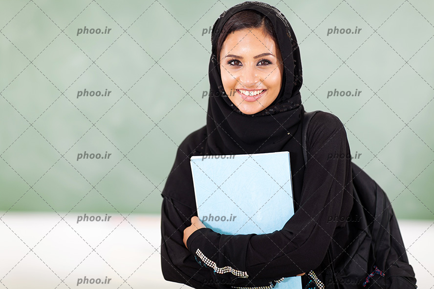 عکس یک خانم با حجاب با لباس و شال مشکی که کوله پشتی دارد و یک جزوه در دستش گرفته است