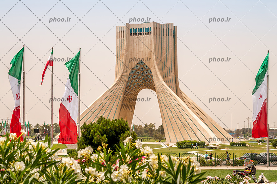 عکس برج آزادی و آسمانی ابری و سبزه های اطراف آن و پرچم ایران