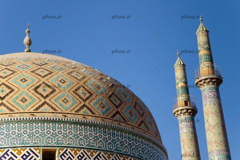 عکس از گنبد و گل دسته های یک مسجد با کاشی های رنگی به شکل لوزی در مقابل آسمان آبی