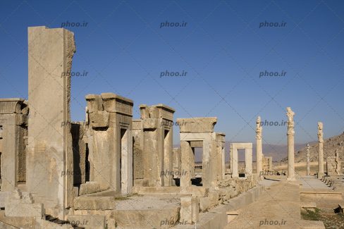 عکس تخت جمشید که در آن دروازه ها و ستون هایی که از سنگ ساخته شده است دیده می شود و آسما آبی