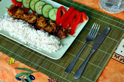 عکس یک دیس غذا از کباب و برنج و گوجه و خیار های تیکه شده در ظرف و کارد و چنگال کنار آن