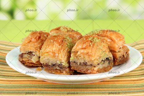 عکس چند شیرینی در یک بشقاب سفید و تزئین شده با مغز پسته و پس زمینه سبز