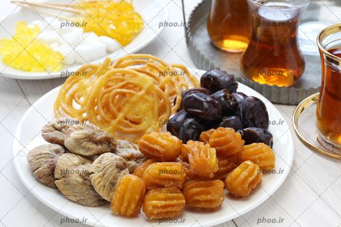 عکس یک ظرف از زولبیا ، بامیه و خرما ، انجیر در کنار چای با نبات و قند