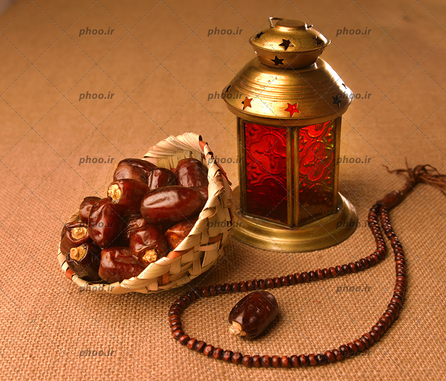 عکس مقداری خرما در یک کاسه حصیری در کنار یک فانوس طلایی با شیشه های قرمز و یک تسبیح روی زمین