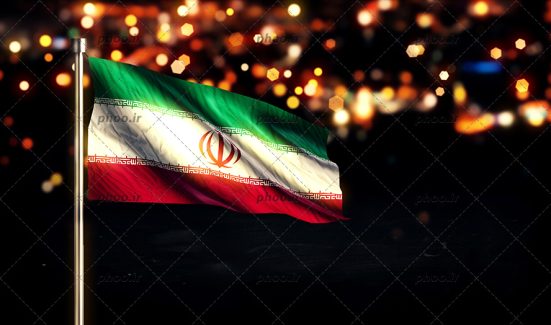 عکس پرچم برافراشته ایران در یک شب تاریک با نور های چراغی که از شهر پیداست