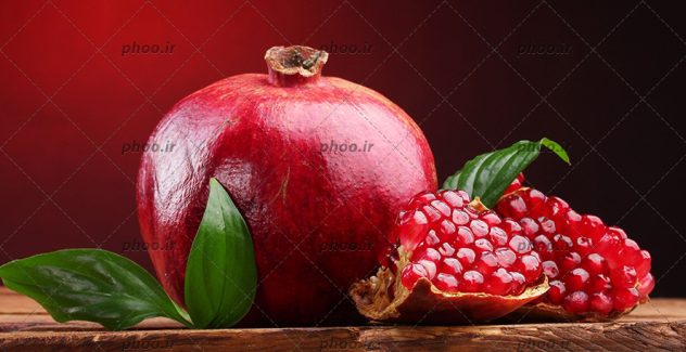 انار به همراه انار نصف شده بر روی میز چوبی با پیش زمینه قرمز و برگ سبز انار