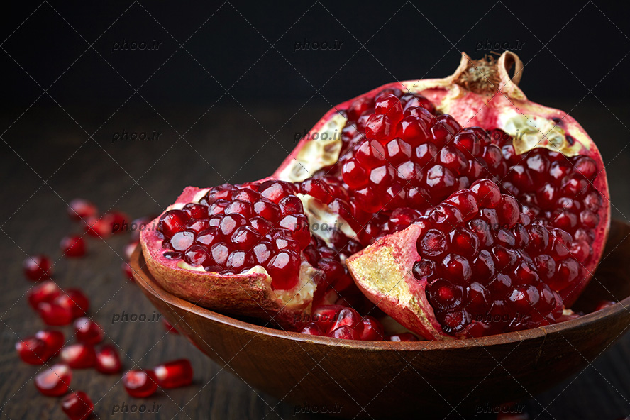 انار نصف شده در ظرف بر روی میز چوبی با دانه های قرمز انار در اطراف