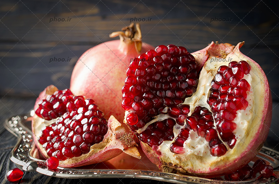 انار به همراه انار دانه شده در ظرف سینی استیل و دانه انار در اطراف بر روی میز چوبی