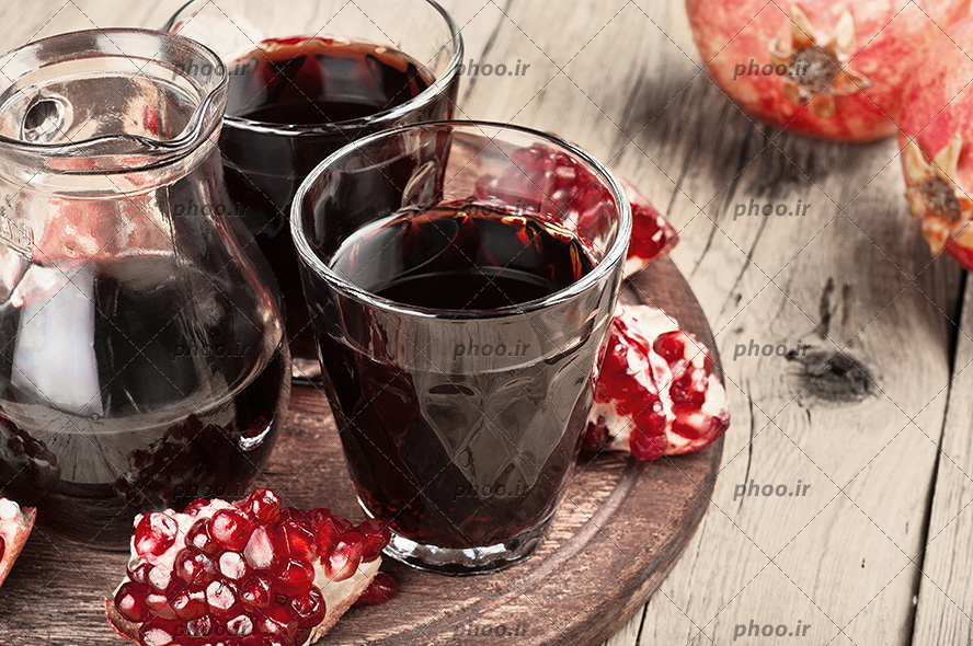 انار نصف شده به همراه آب انار در پارچ و دو لیوان شیشه ای در سینی چوبی بر روی میز چوبی