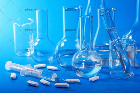 عکس با کیفیت شیشه آلات آزمایشگاهی و قرص و آمپول در کنار یکدیگر در پس زمینه آبی