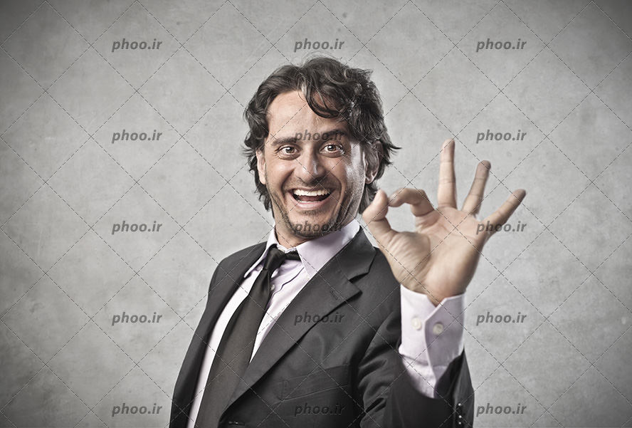 عکس با کیفیت مرد با کت و شلوار مشکی و لبخند به لب در حال نشان دادن علامت خوب با استفاده از انگشتان خود
