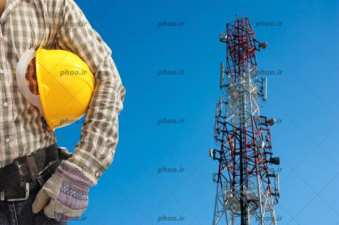 عکس با کیفیت مرد ایستاده با کلاه ایمنی زرد در بغل و دکل برقی در کنارش