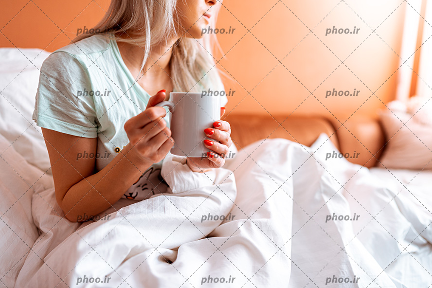 عکس خانم لیوان سفید به دست در تخت خواب سفید در اتاق نارنجی هنگام از خواب پاشدن