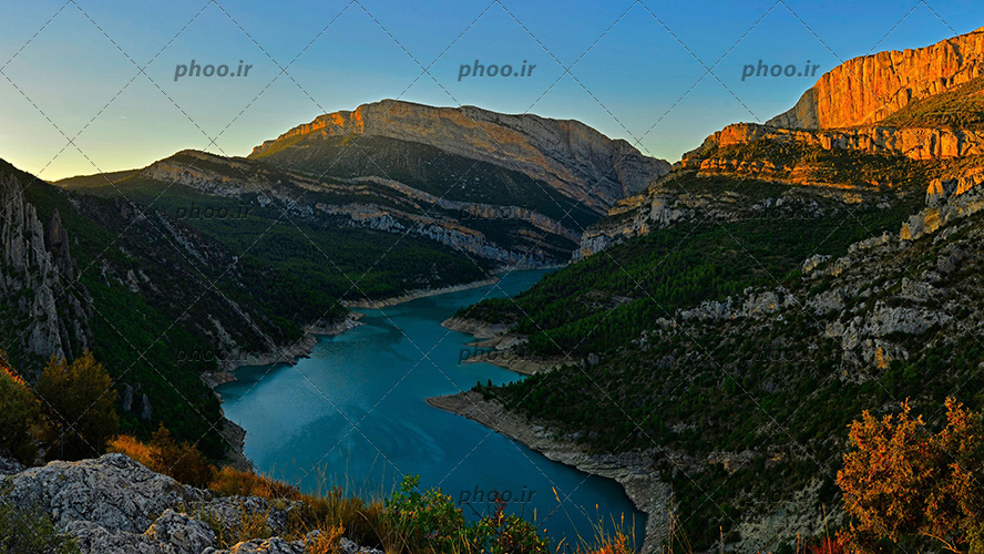 عکس طبیعت زیبا کوهستانی و صخره ای با دریاچه یا رودخانه و درختان کاج و آسمان آبی