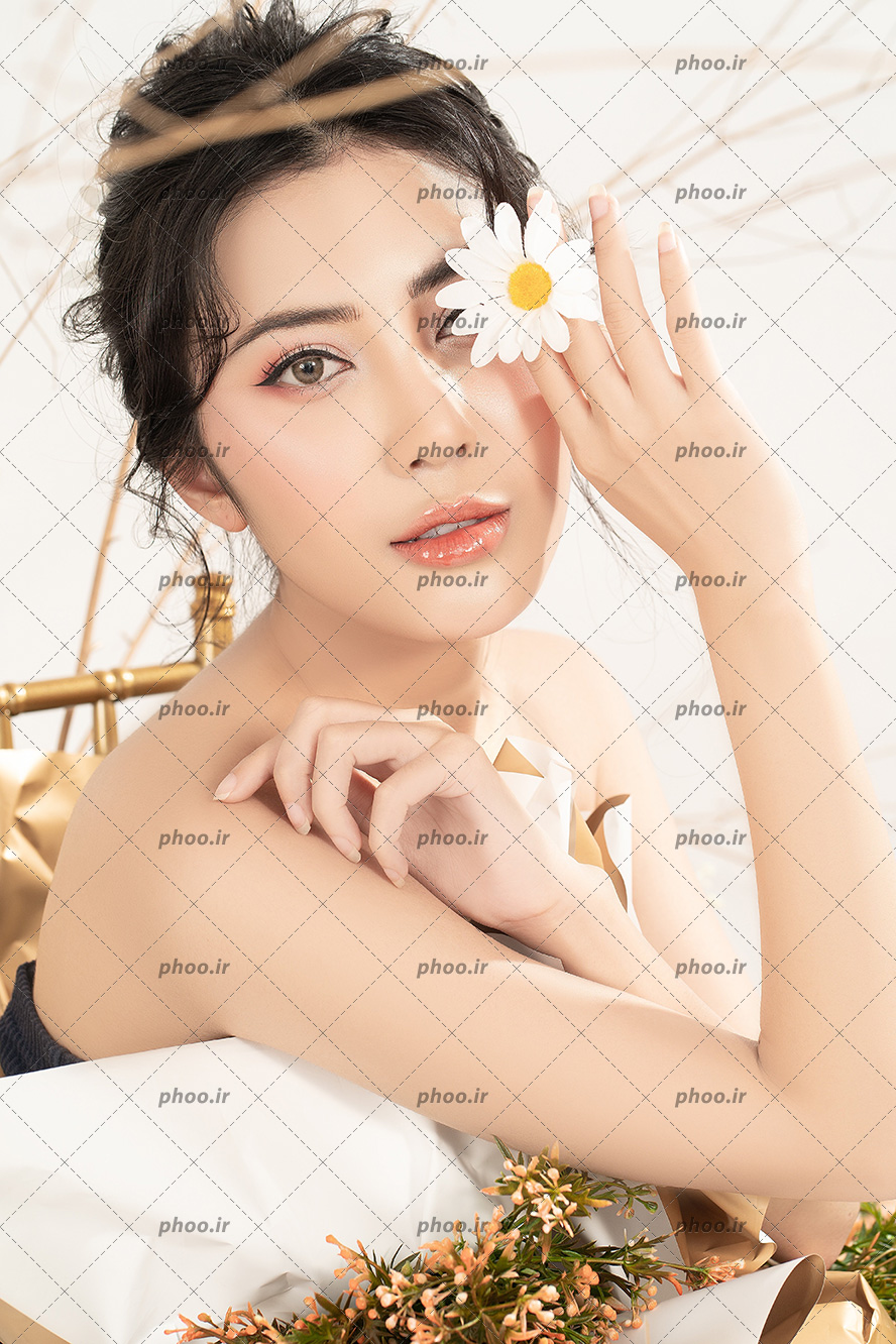 عکس مدل دختر نشسته در تخت و گل را جلوی چشم خود گرفته با زمینه سفید