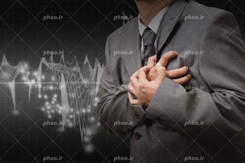 عکس با کیفیت مرد با کت شلوار خاکستری در حال گرفتن قلب خود با دست و در بک گراند مشکی و ضربان قلب در زمینه آن