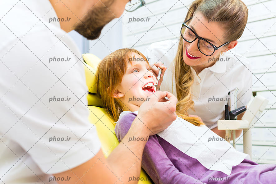 عکس با کیفیت کودک زیبا دراز کشیده بر روی صندلی مخصوص دندانپزشکی و دو پزشک در حال معاینه دندان هایش