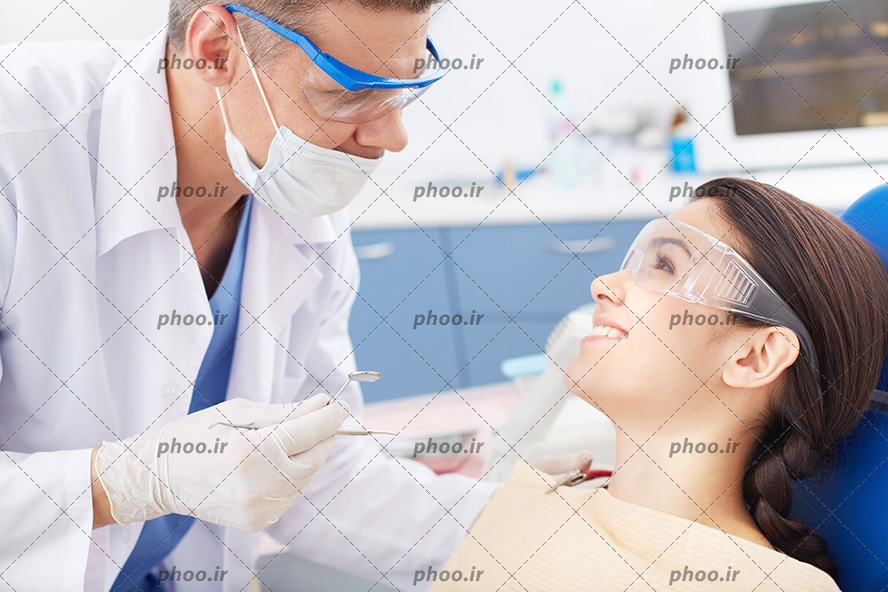 عکس با کیفیت پزشک با روپوش سفید و عینک محافظ در حال صحبت با بیمار دراز کشیده بر روی صندلی و در حال لبخند زدن و عینک ماحفظ بر روی صورتش