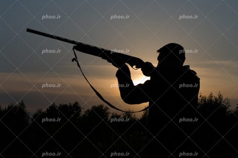 عکس با کیفیت اسلحه شکاری در دست مرد شکارچی و خورشید در حال غروب
