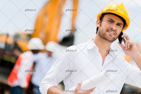 عکس با کیفیت مهندس ساختمان با پیرهن سفید و کلاه ایمنی به رنگ زرد بر روی سر در حال مکالمه با تلفن