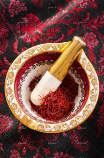 عکس با کیفیت زعفران های تازه داخل هاون طلایی سفید با طرح زیبا و قرار گرفته بر روی پارچه ای زیبا به رنگ مشکی قرمز و طرح های اسلیمی بر روی پارچه