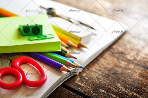 عکس با کیفیت قیچی و تراش و مداد های رنگی و پرگار و نوت بوک بر روی دفتر با صفحات باز