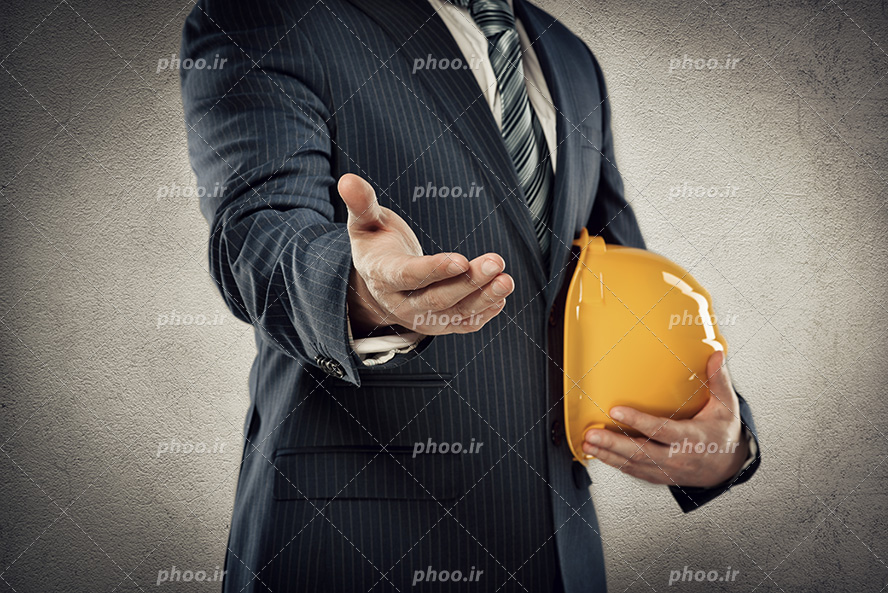 عکس با کیفیت زیبا مرد ایستاده با کت و شلوار و کلاه ایمنی در دست در حال اشاره با دست