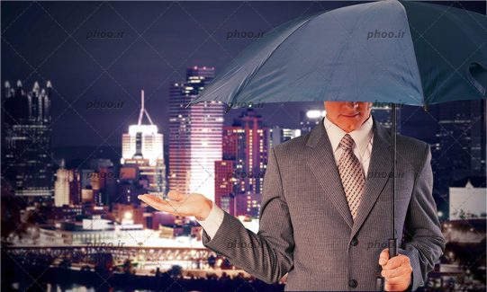 عکس تاج با کت و چتر در دست در حال چک کردن باران در شب در پس زمینه ساختمان های شهر