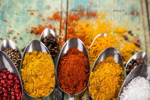 عکس ادویه های متفاوت در قاشق های فلزی از نمای نزدیک در پس زمینه چوبی