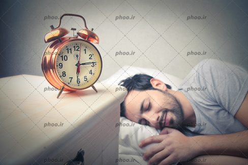 عکس مرد خوابیده در رختخواب و ساعت رومیزی زنگ دار روی میز
