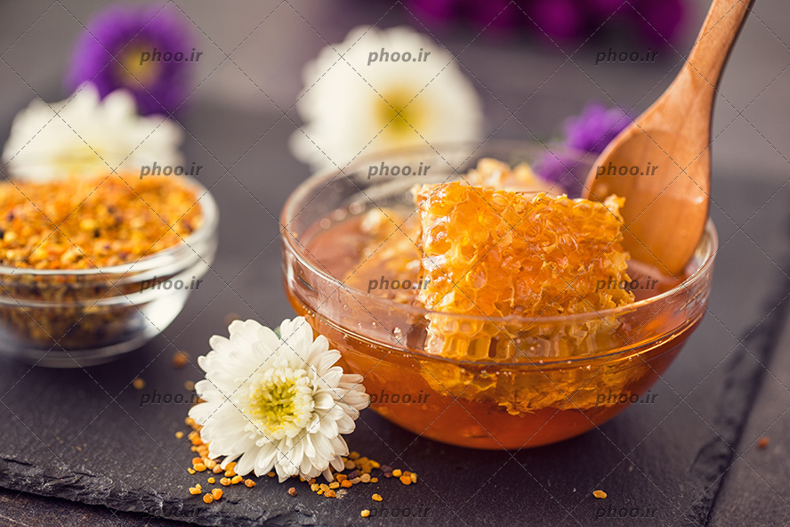 عکس عسل طبیعی در کاسه شیشه ای و قاشق چوبی در آن در کنار گل های سفید و بنفش پس زمینه مشکی