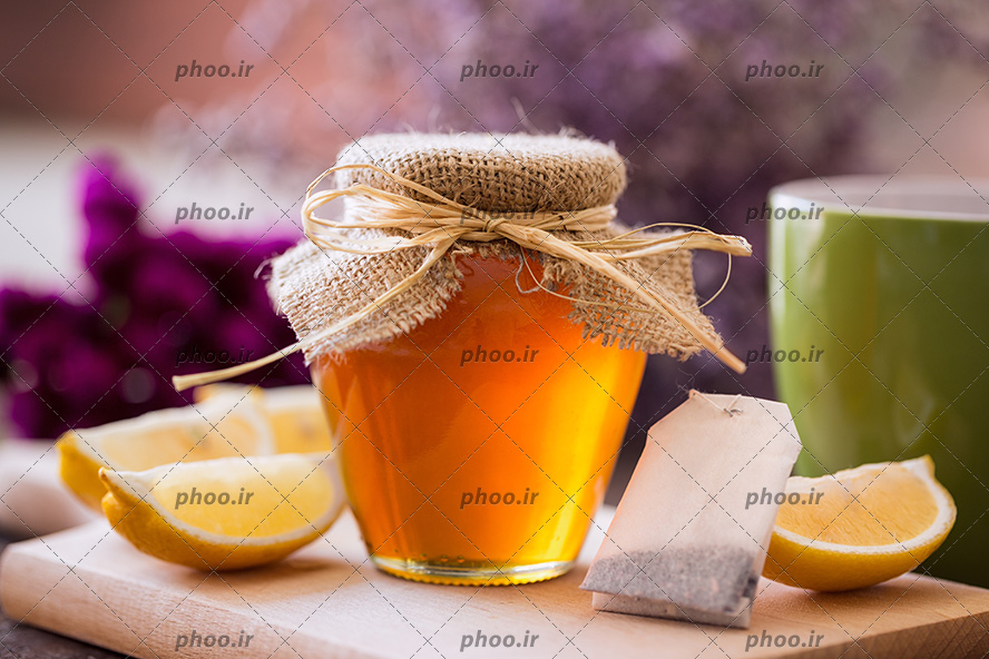 عکس ظرف عسل در کنار چای کیسه ای و برش های لیمو روی تخته چوبی