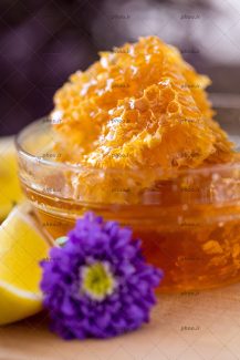 عکس عسل طبیعی در کاسه شیشه ای در کنار برش های لیمو و یک گل بنفش