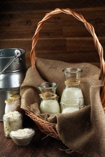 عکس لبنیات مقوی در ظرف های شیشه ای روی زمین و داخل سبد در کنار سطل فلزی در پس زمینه چوبی