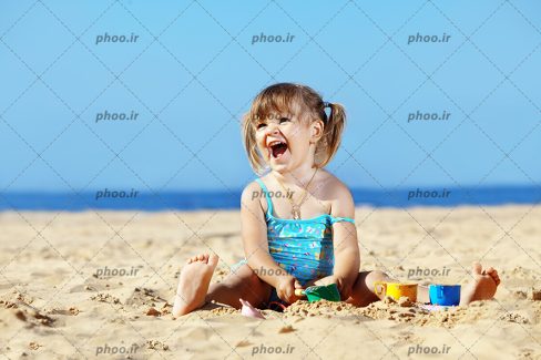 عکس با کیفیت دختر بچه شاد در حال بازی با ماسه ها در پس زمینه آبی